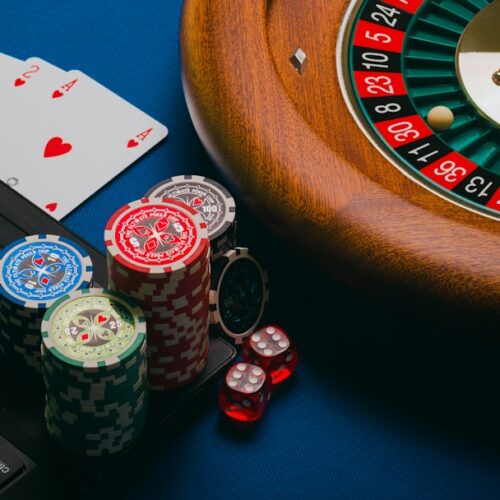 Close-up of poker chips, cards, and a roulette wheel on a blue surface with a laptop partially visible.