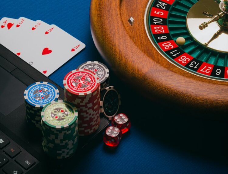 Close-up of poker chips, cards, and a roulette wheel on a blue surface with a laptop partially visible.