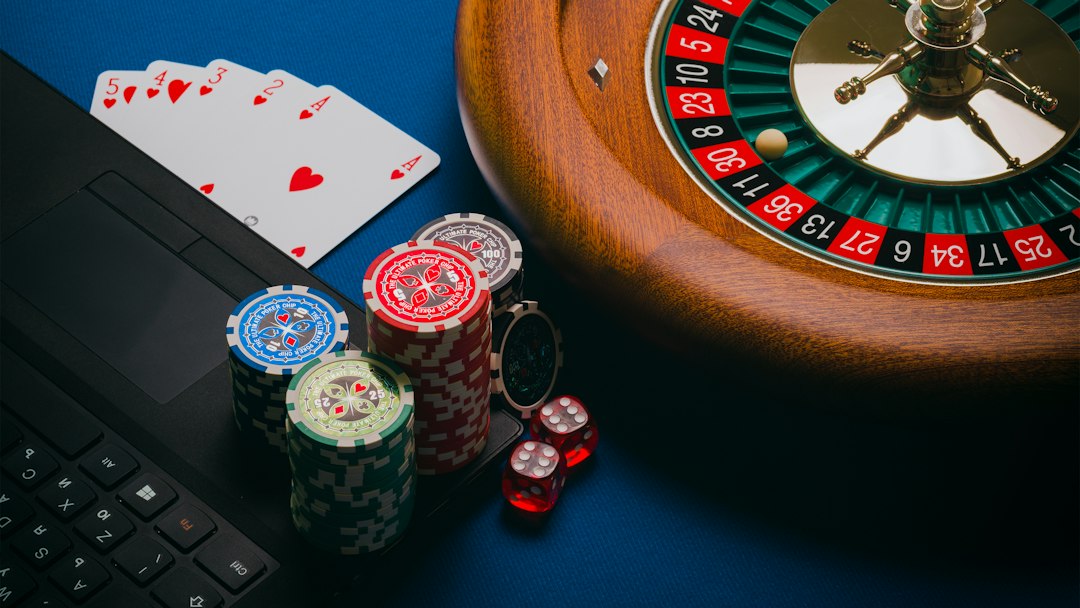 Close-up of poker chips, cards, and a roulette wheel on a blue surface with a laptop partially visible.